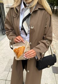 Γυναίκα με μπεζ παλτό τραντσκότ που κρατά ένα κρουασάν τυλιγμένο σε χαρτί, φοράει μαύρο και λευκό κασκόλ και κρατά μια μαύρη τσάντα με καπιτονέ σχέδιο.