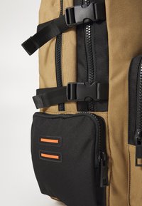 Tan backpack with black accents, featuring multiple zippers and buckles. Front pocket has two horizontal orange stripes. Durable fabric texture.