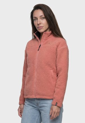 Frau trägt eine lachsrosa strukturierte Fleecejacke mit Reißverschluss vorne und hellblaue Jeans, steht vor einem einfarbigen hellen Hintergrund.