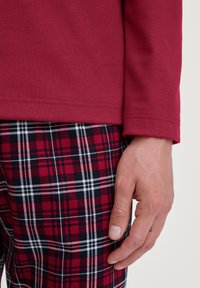 Rode overhemd met lange mouwen, gecombineerd met zwart, rood en witte geruite broek. De materialen lijken zacht met een gladde textuur. Een hand rust op de broek.