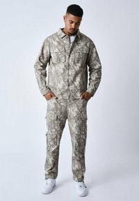 Camo-patroon lichte katoenen jas en cargo-broek set in gedempt beige en grijs, met meerdere zakken en knoopsluitingen. Witte sneakers.