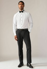 Witte overhemdd met zwarte knopen en een zwarte vlinderdas, gecombineerd met op maat gemaakte zwarte broek en zwarte nette schoenen. Soepele, formele stijl.