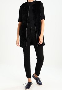 Svart konstläderväst med ståkrage och framsidans dragkedja, kombinerad med en svart topp, åtsittande svarta byxor och mörkblå snörskor.