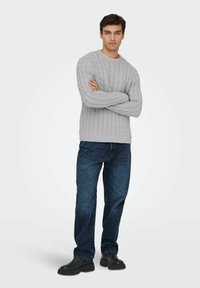 Un jeune homme se tenant debout, les bras croisés, vêtu d'un pull en laine gris à tresses, d'un jean bleu foncé et de bottes noires, sur un fond uni.