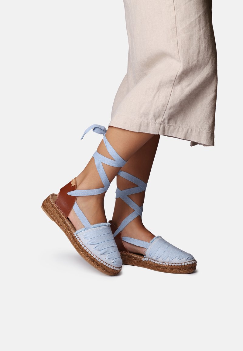 Voeten met lichtblauwe espadrilles sandalen met enkelstriklinten en een bruine hielriem, gecombineerd met beige cropped broek.