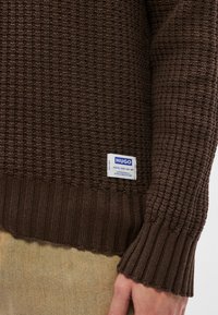 Barna kötött pulóver texturált mintával és bordázott mandzsettákkal. Bal alsó oldalon kék-fehér "HUGO" feliratos címkével.