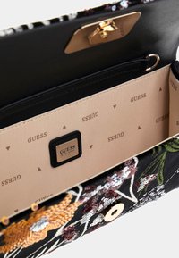 Zwarte geborduurde handtas met bloemmotieven, gouden hardware en een beige interieur met het GUESS-logo in een herhalend patroon.