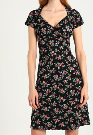 Zwarte jurk met korte mouwen en een V-hals met een meerkleurig klein bloemenpatroon, gedragen door een staande vrouw.