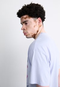 Lyse lilla bomulds T-shirt med grafiske tryk i forskellige farver. Modellen har kort, krøllet hår og synlige hudmærker.