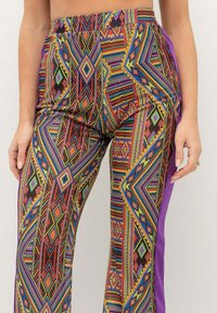 Pantaloni colorati a fantasia con un design geometrico in rosso, blu, verde e giallo, con un pannello laterale solido viola e una cintura elastica.