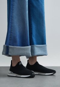 Ténis pretos de malha sem atacadores com parte superior texturizada, com um detalhe prateado na sola. Combinados com jeans de ganga azul de perna larga.