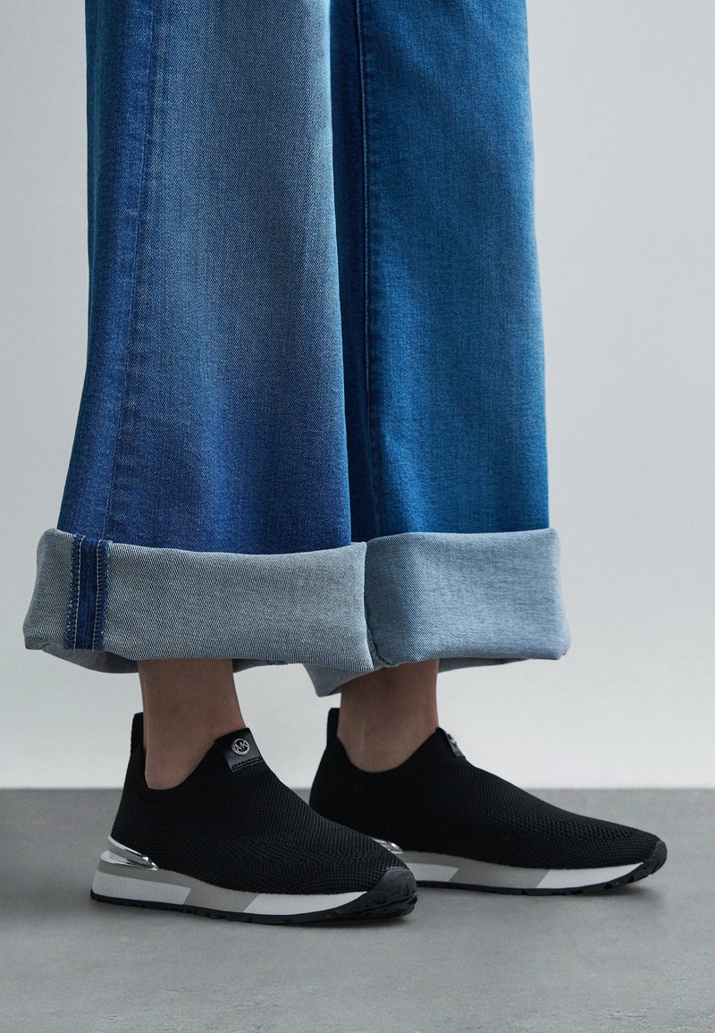 Ténis pretos de malha sem atacadores com parte superior texturizada, com um detalhe prateado na sola. Combinados com jeans de ganga azul de perna larga.