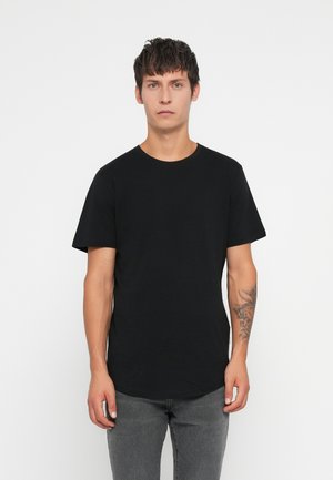 Junger Mann mit dunklem Haar, der ein schlichtes schwarzes Kurzarm-T-Shirt und graue Jeans trägt, vor einem schlichten weißen Hintergrund steht.