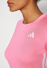 Rózsaszín sporttop kerek nyakkivágással, testhez simuló fazonnal és fehér logó részletekkel. Az anyag simának és rugalmasnak tűnik.