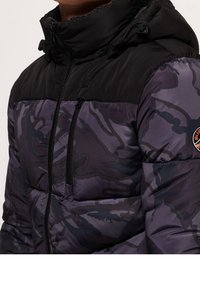 Puffjacka med en svart övre del och lila camouflage-mönster. Har dragkedjestängning, hög krage och en dragkedjeförsedd bröstficka.