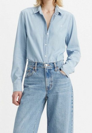 Personne portant une chemise bleu clair boutonnée rentrée dans un jean bleu taille haute et ample, une main dans la poche.
