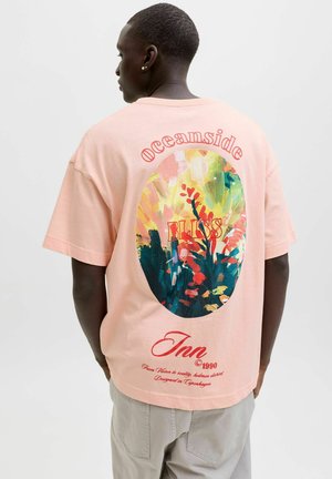 Vīrietis valkā gaiši rozā t-kreklu ar krāsainu ziedu ovālu grafiku un sarkaniem burtiem uz muguras: "oceanside", "BLISS" un "Inn".