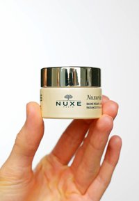 NUXE Nuxuriance Radiance ögonbalsam i en liten, rund glasburk med ett gyllene lock, med en krämfärgad etikett som har text och logotyp.