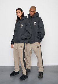 Tmavošedé mikiny na zips s prednými vreckami, béžové nohavice s čiernymi postrannými pruhmi a logom, a čierne tenisky. Jednoduchý dizajn a pohodlný strih.