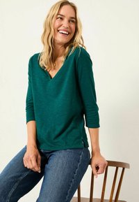 Green v-neck top with three-quarter sleeves, featuring a relaxed fit and side slits, paired with blue denim jeans. Smooth texture.