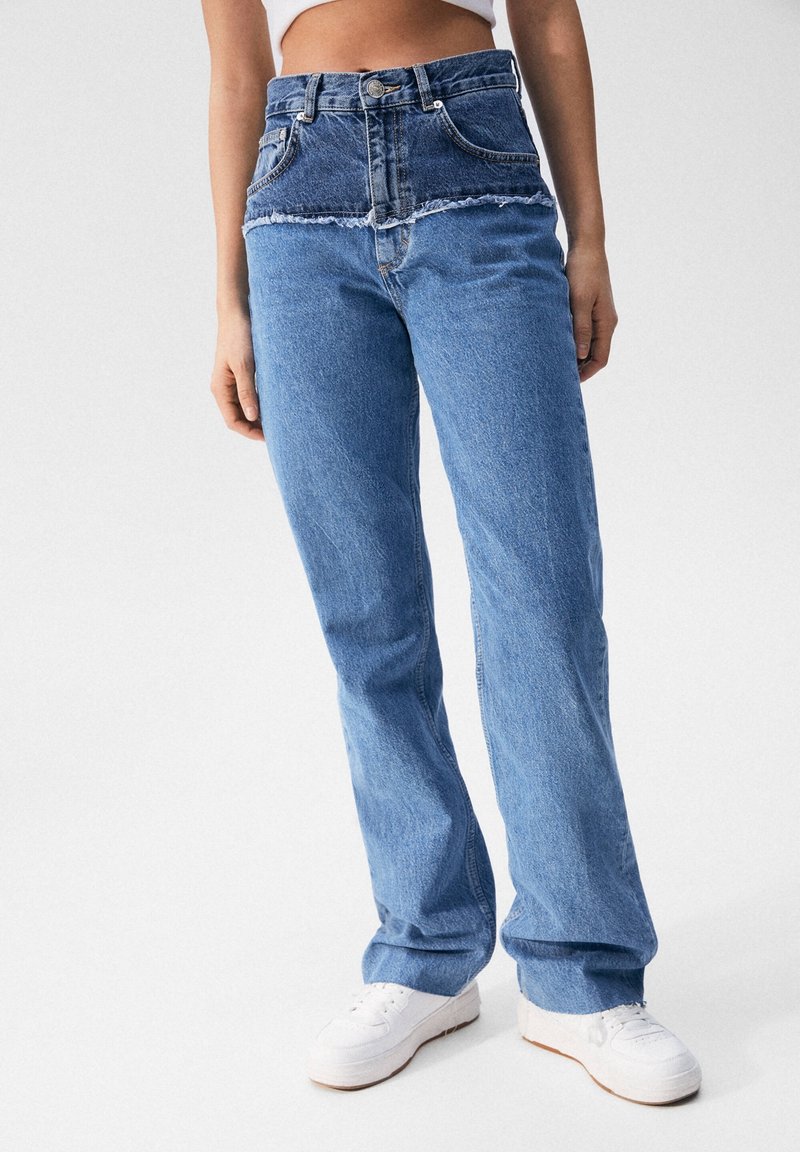 Femme portant un jean bleu bicolore taille haute avec une couture effilochée à la taille et des baskets blanches sur un fond uni.