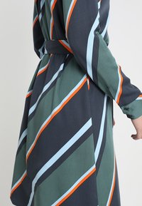 Pitkähihaisen tummanvihreän mekon, jossa on viistosti suuntautuvat vaaleansiniset ja oranssit raidat. Sisältää vyötärölle pujotettavan vyön ja sileän kankaan rakenteen.