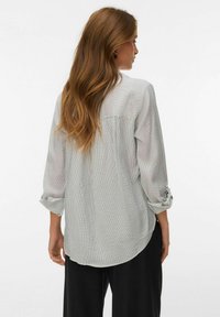 Femme aux longs cheveux bruns portant un haut à manches longues rayé blanc et gris et un pantalon noir, vue de dos avec la tête tournée sur le côté.