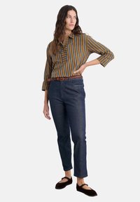 Prugasta košulja s ovratnikom u senfu žutoj i tamnozelenoj boji, uparena s tamnim trapericama. Naglašena pletenim smeškim remenom i tamnosmešnim cipelama.