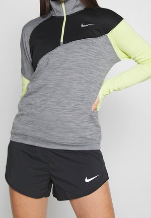 Kobieta ubrana w szarą i czarną koszulkę Nike z długim rękawem, neonowo żółtymi rękawami i czarne spodenki Nike, stojąca z jedną ręką na biodrze.