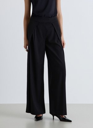 Donna che indossa pantaloni neri a vita alta e a gamba larga e tacchi alti neri con punta a punta, in piedi davanti a uno sfondo chiaro e uniforme.