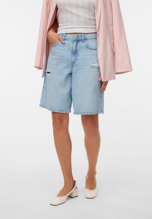 Femme portant un short en jean déchiré bleu clair longueur genou, un top blanc côtelé, un blazer rose et des chaussures crème à talons blocs, debout.