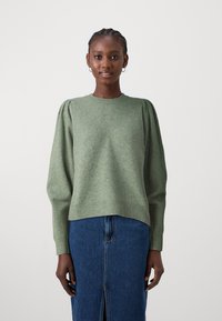 Zeleno pleteno pulover s puhastimi rokavi, z teksturiranim diamantom vzorcem na sprednji strani, rebrastimi manšetami in robom. Kombiniran z denim krilom.