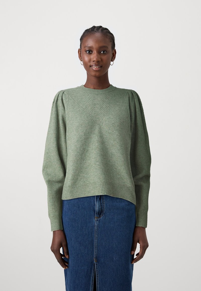 Zeleno pleteno pulover s puhastimi rokavi, z teksturiranim diamantom vzorcem na sprednji strani, rebrastimi manšetami in robom. Kombiniran z denim krilom.