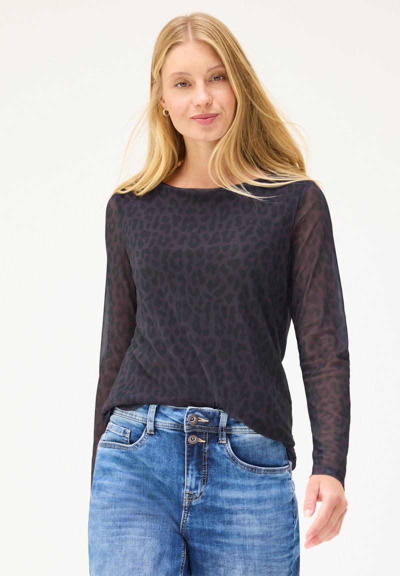 Femme blonde portant un haut noir transparent à manches longues avec un subtil imprimé léopard et un jean en denim bleu, debout devant un fond uni.