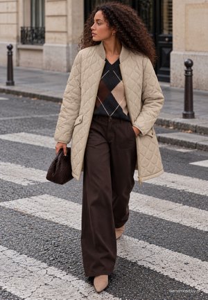Une femme traverse une rue en ville, vêtue d’un manteau matelassé beige, d’un pull à motifs et d’un pantalon marron, avec un sac à main foncé.