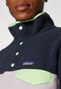 Fleece-pullover med marineblå kropp, lysegrå nederdel og grønn krage. Har trykknapper og et Patagonia-logo på brystlommen.