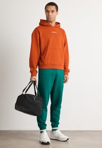 Felpa di cotone arancione con tasca frontale, abbinata a pantaloni felpati verdi. Una borsa a tracolla nera e scarpe da ginnastica bianche completano l'outfit.