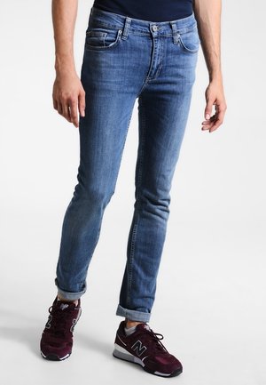 Man draagt slim-fit blauwe spijkerbroek met opgerolde pijpen en donkerrode New Balance sneakers, staand op een witte achtergrond.