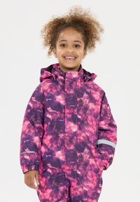 Smilende barn med krøllet hår iført en vandtæt jakke og bukser med lilla og pink sky-mønster samt reflekterende stripe på ærmet.