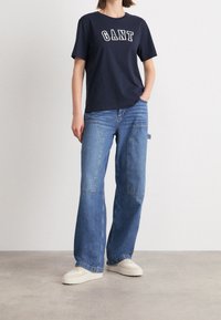 Marineblauwe katoenen t-shirt met wit "GANT" logo. Lichtblauwe denim broek met brede pijpen en een vervaagde afwerking, met zijzakken. Beige sneakers.