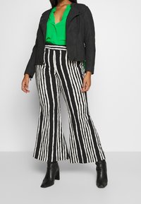 Giacca motociclista in pelle nera testurizzata, blusa verde e pantaloni a zampa neri e bianchi a righe con linee verticali audaci. Stivaletti neri con tacco.