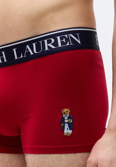 Κοντινό πλάνο ενός ατόμου που φοράει κόκκινα ανδρικά εσώρουχα μποξέρ με μπλε σκούρο ελαστικό περίζωμα στη μέση με την επιγραφή "LAUREN" και ένα μικρό κεντητό αρκουδάκι με ρόμπα.