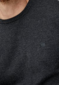 Gros plan sur une personne portant un pull en tricot gris foncé avec un petit logo discret sur la poitrine près de l'encolure.