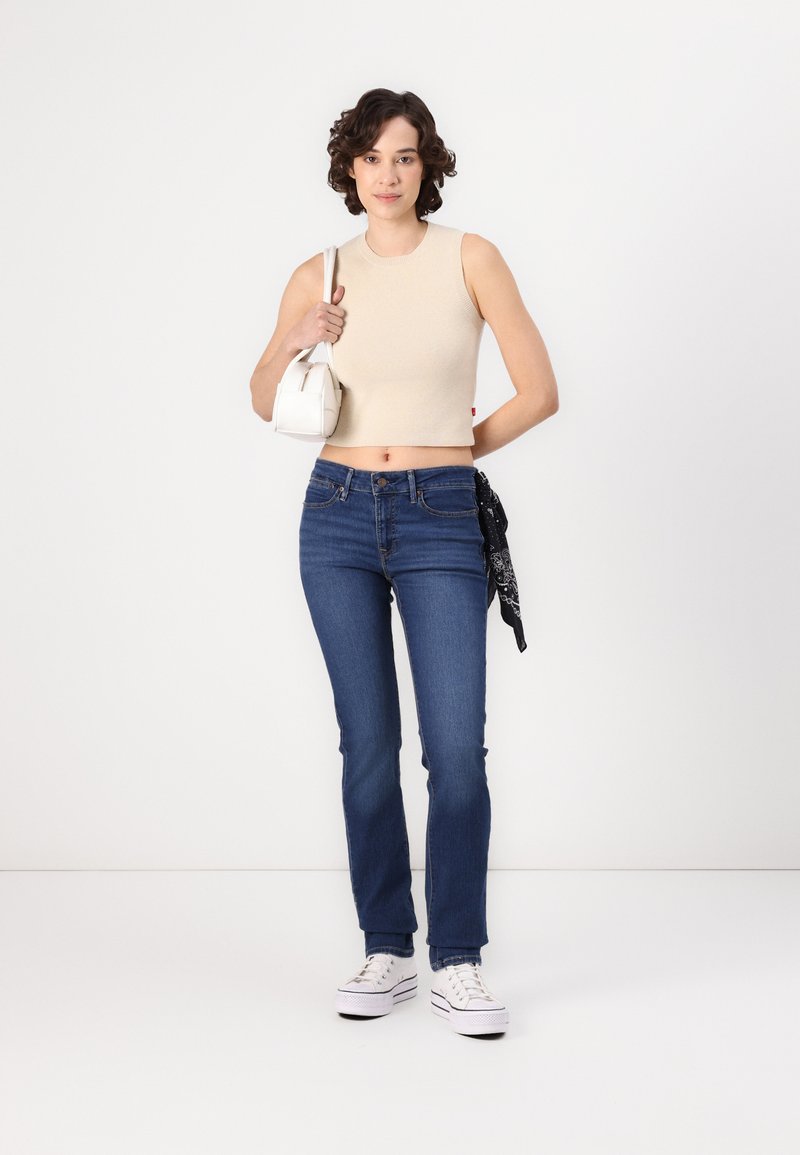 Top corto de canalé beige, jeans azul oscuro, zapatillas blancas y bolso blanco pequeño con un pañuelo negro atado a la cintura.