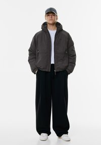 Pufferjack in donkergrijs met een capuchon, met voorzakken met rits, gecombineerd met losse zwarte broek en witte sneakers, tegen een effen achtergrond.
