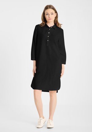 Vrouw staat en draagt een zwarte knielange jurk met knopen, lange mouwen en witte sneakers tegen een effen witte achtergrond.