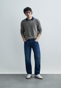 Gray knit sweater with navy collar, blue denim jeans, and white sneakers with brown soles. Model stands with hands in pockets against a neutral background.