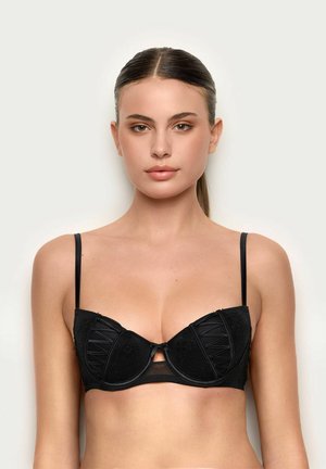 Ung kvinde med glat brunt hår trukket tilbage iført en sort blonde-bh med bøjle, mod en ensartet lys baggrund.