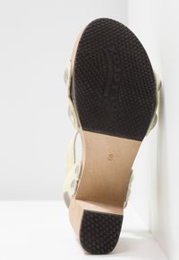 Semelle de sandale avec talon et avant-pied en caoutchouc noir texturé, semelle intermédiaire en bois beige, et brides de couleur claire ornées de décorations circulaires.