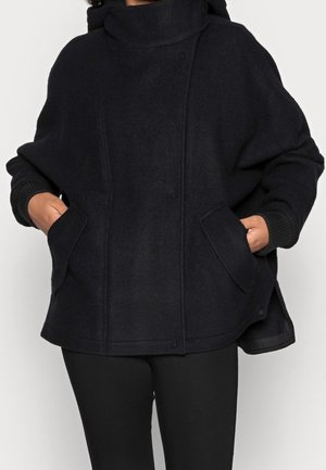Personne portant un manteau en laine noir oversize avec un col montant, des fentes latérales et les mains dans les poches avant, associé à un pantalon noir.